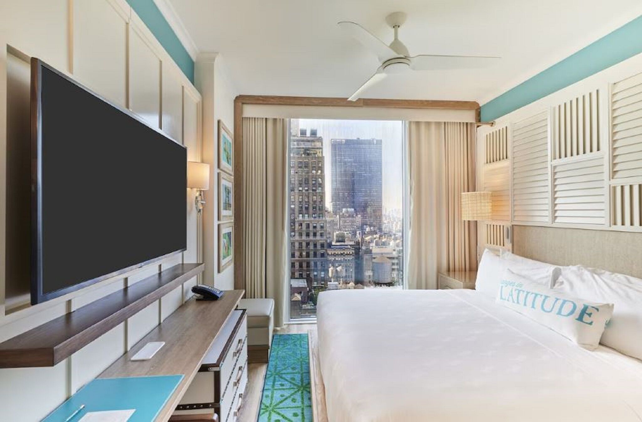 A hotel room with a large bed, a flat-screen TV, a desk with a lamp, and a view of the cityscape.