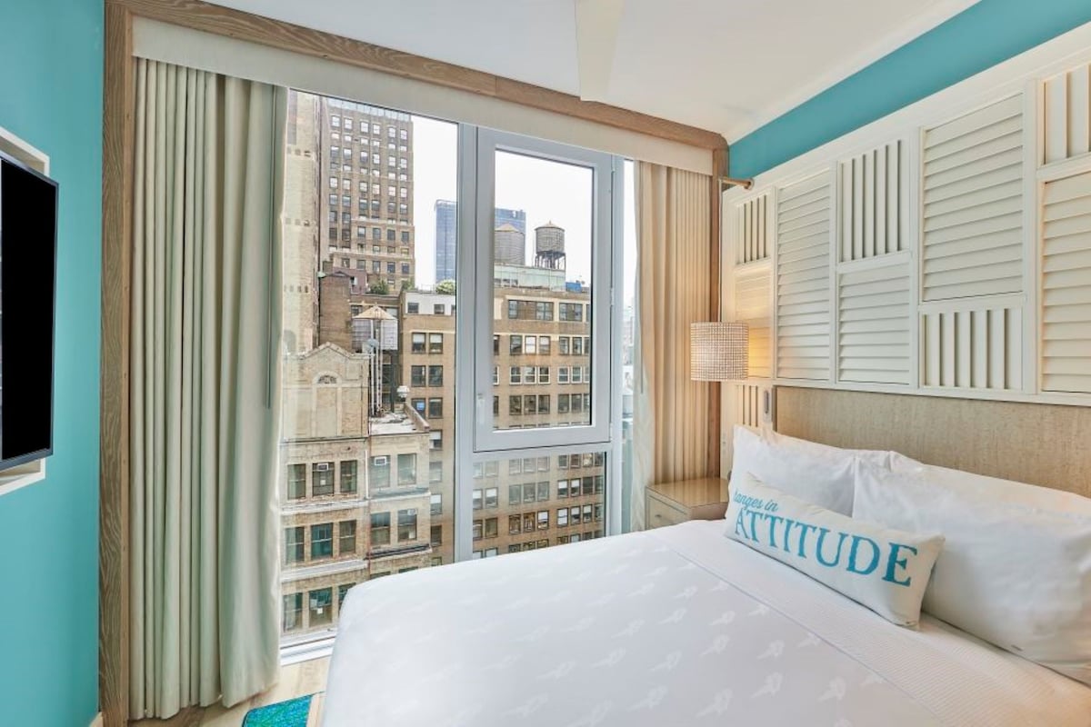 A hotel room with a large bed, a view of city buildings, a TV, and a window with curtains.