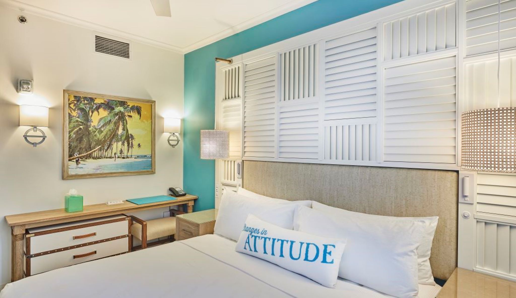 A hotel room with a bed, a desk, a chair, a painting, and a wall with shutters.