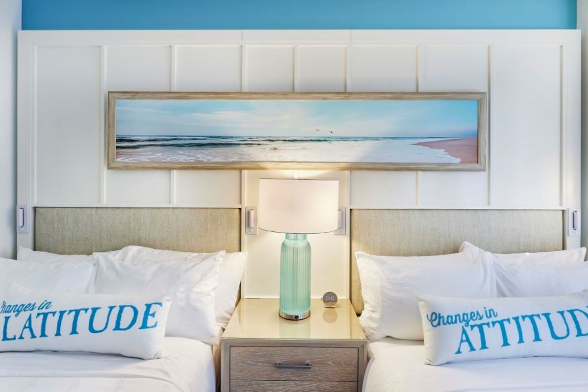 A hotel room with two beds, a nightstand with a lamp, and a painting of a beach scene.