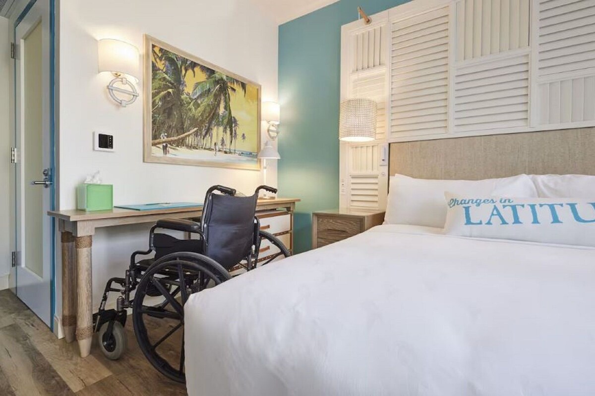A hotel room with a bed, a desk, a wheelchair, a painting, and a wall-mounted light fixture.