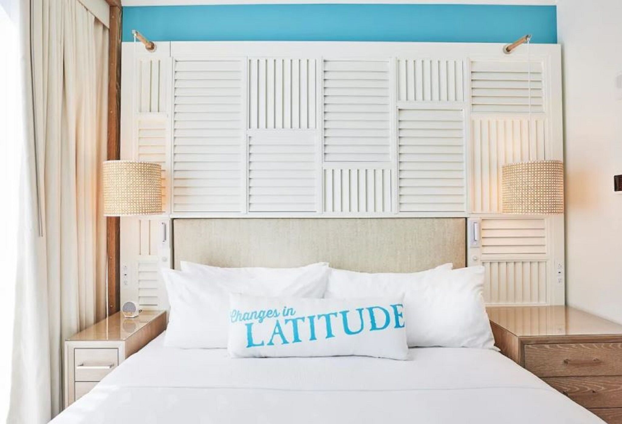 A bed with a white headboard, a pillow with 'Changes in LATITUDE' text, and two bedside lamps.
