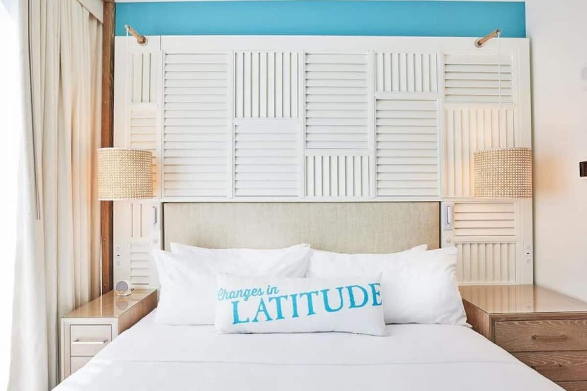 A bed with a white headboard, a pillow with 'Changes in LATITUDE' text, and two bedside lamps.