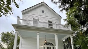 Une maison blanche à deux étages, dotée d’un porche à l’avant, d’un escalier et d’un petit jardin avec des arbustes et un palmier.