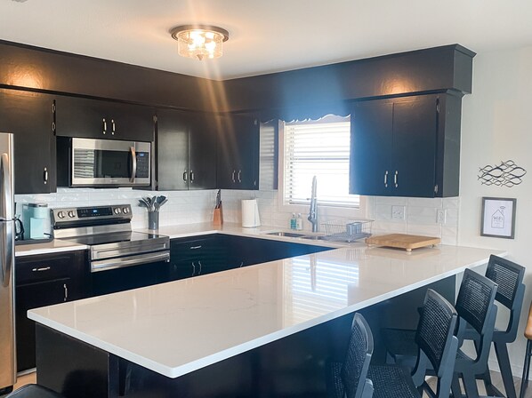 A modern kitchen with dark cabinets, stainless steel appliances, a white countertop, and a window providing natural light. - Sunny Family Friendly House 1500 sqft ~ Oversized Covered Deck ~ .5 m to Beach (Port O'Connor)