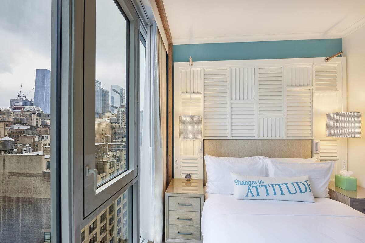 A hotel room with a city view, a bed with a pillow labeled "Changes in Attitude," and a nightstand with a lamp.