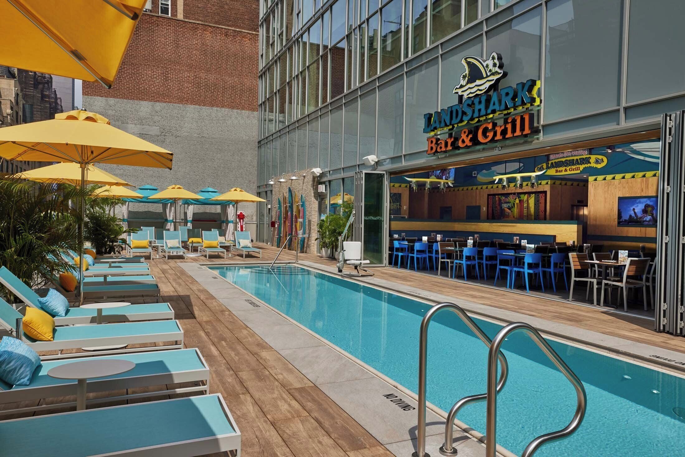Zona de piscina con sillones de descanso y sombrillas, un bar y restaurante llamado 'Landshark', y un edificio de ladrillo al fondo.