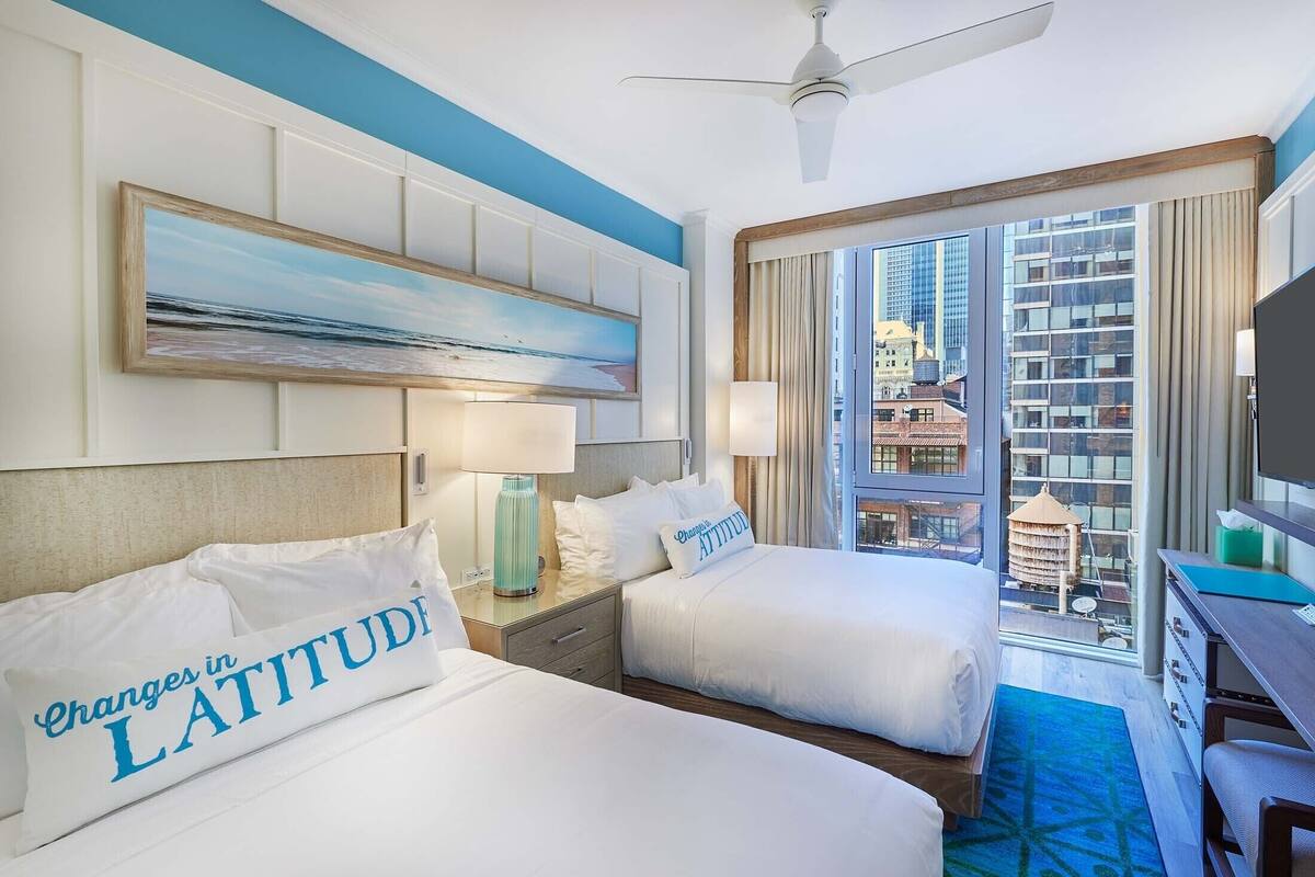 A hotel room with two beds, a large window with a city view, a ceiling fan, and a painting on the wall.