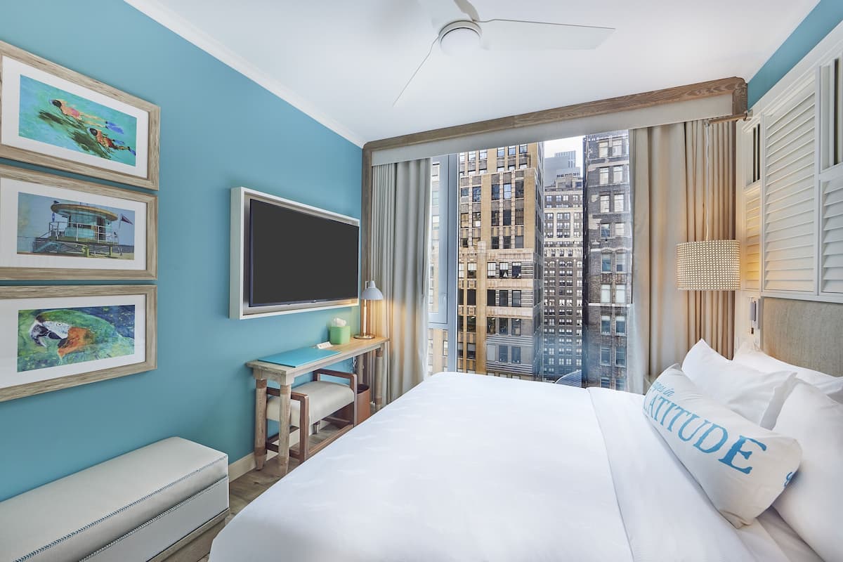 A hotel room with a bed, a desk, a chair, a TV, and a window with a city view.
