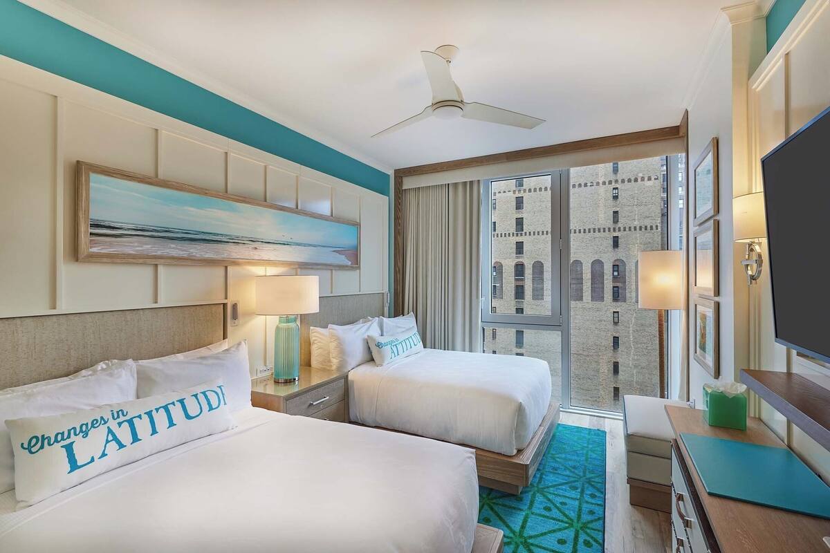 A hotel room with two beds, a ceiling fan, a large window with curtains, and a view of a building.