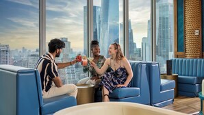 A modern lounge with blue seating, large windows offering a city view, and a bar area with a drink being served. - Margaritaville Resort Times Square (New York)
