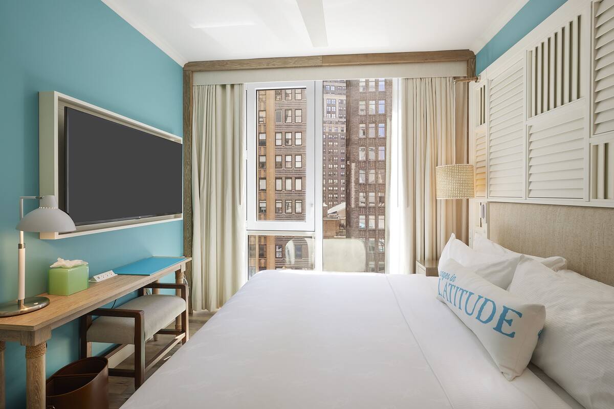 A hotel room with a large bed, a desk with a computer, a chair, a window with curtains, and a view of buildings.