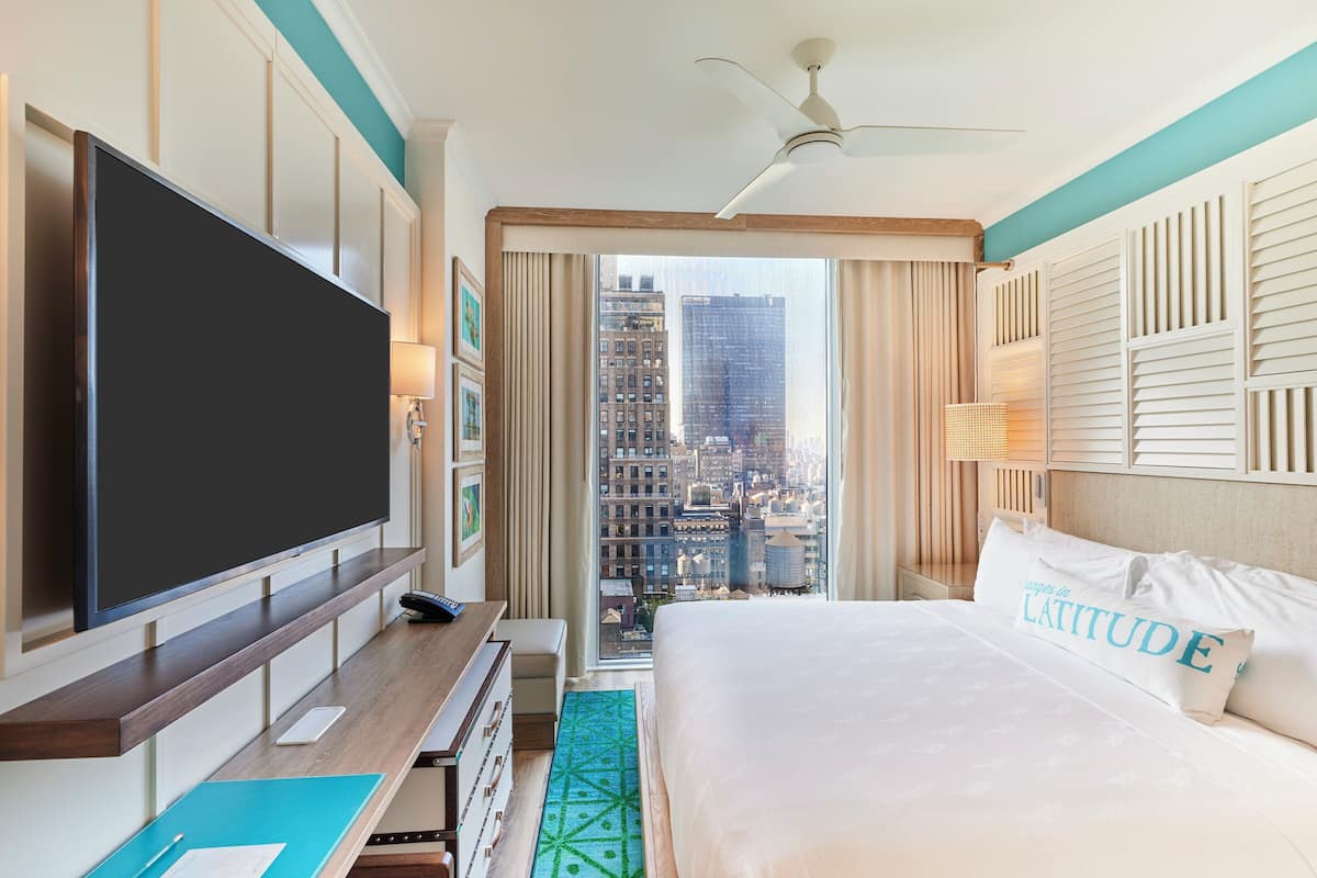 A hotel room with a large bed, a flat-screen TV, a desk with a laptop, and a view of a cityscape through a large window.