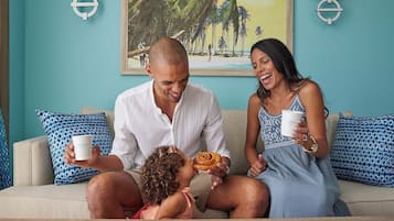 Una familia de tres en un sofá color beige con cojines estampados, un cuadro de playa tropical en la pared y una lámpara a cada lado.