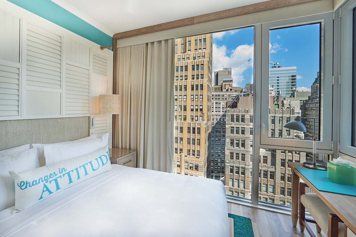 A hotel room with a large bed, a view of the cityscape through floor-to-ceiling windows, a desk with a lamp, and a view of the cityscape through the windows.