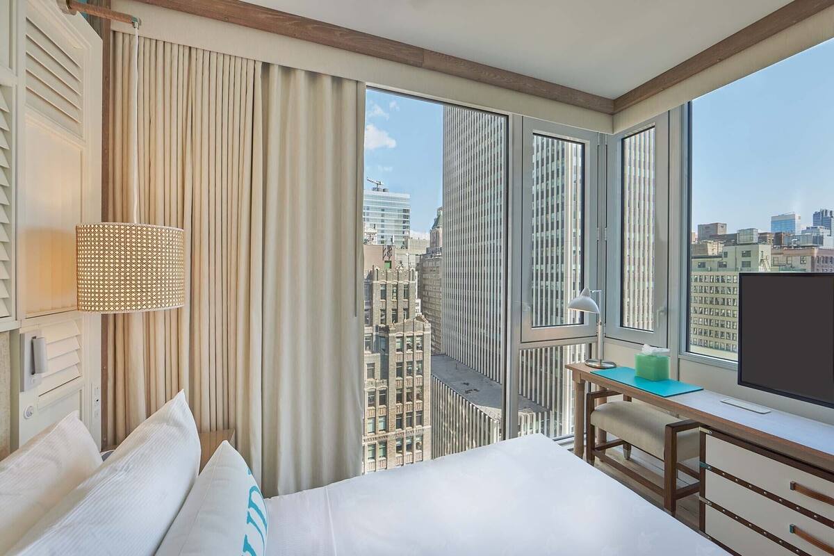 A hotel room with a bed, a desk, a TV, and a large window offering a city view.