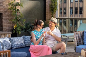 A couple sitting on a blue sofa with patterned cushions, holding drinks, in an outdoor setting with a brick wall and plants in the background.