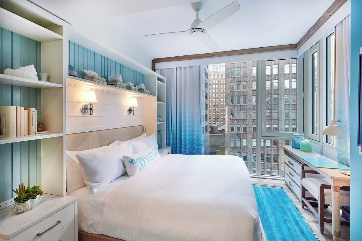 A modern bedroom with a large bed, built-in shelving, a desk, and a city view.