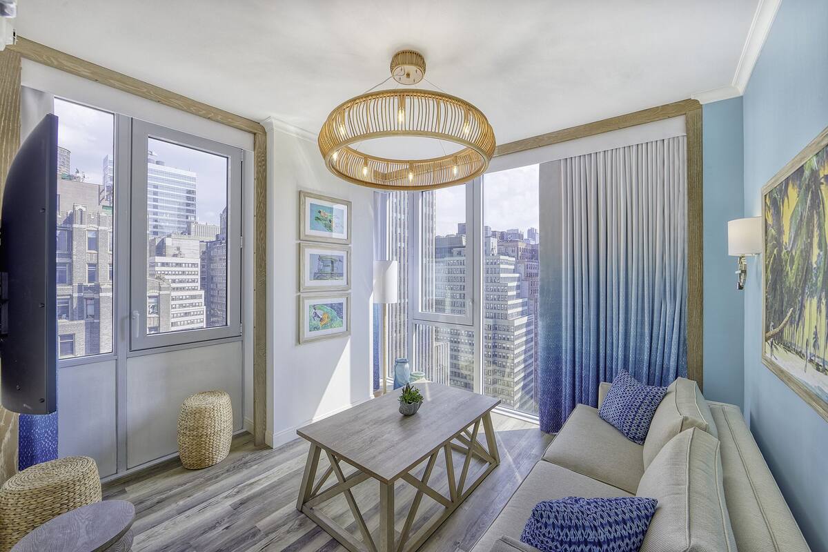 A modern living room with a large window, a wooden table, a sofa, and a hanging light fixture.