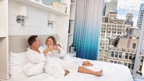 A couple in a hotel room with a city view, a bed with white linens, and a nightstand with a bottle. - Margaritaville Resort Times Square (New York)