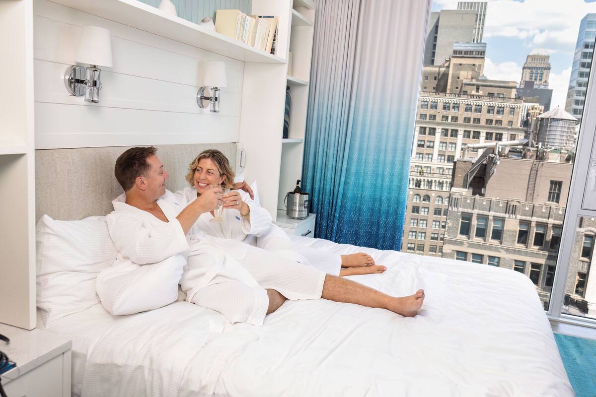 A couple in a hotel room with a city view, a bed with white linens, and a nightstand with a bottle.