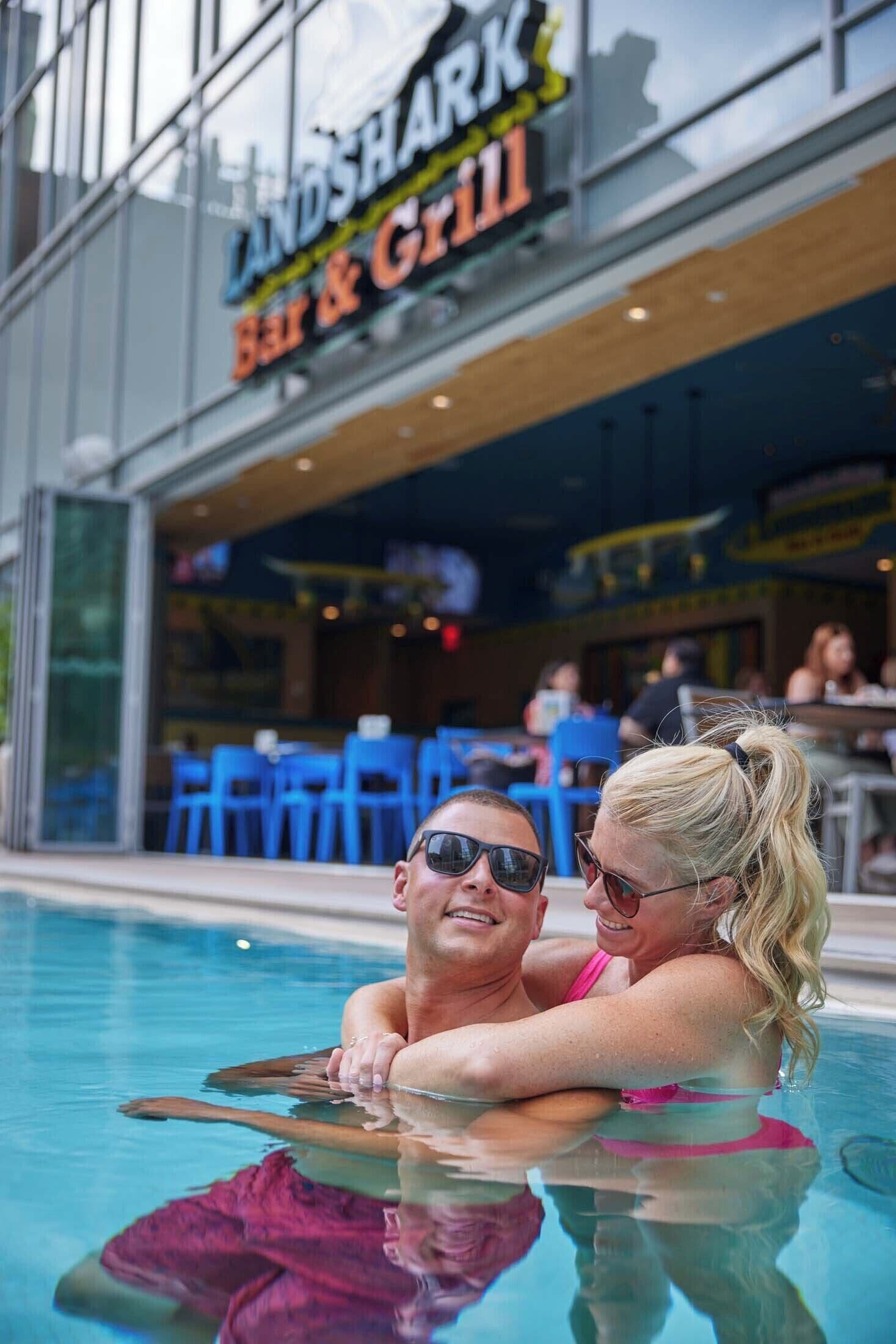 Un couple se détend dans une piscine, avec un bar et un espace barbecue en arrière-plan.