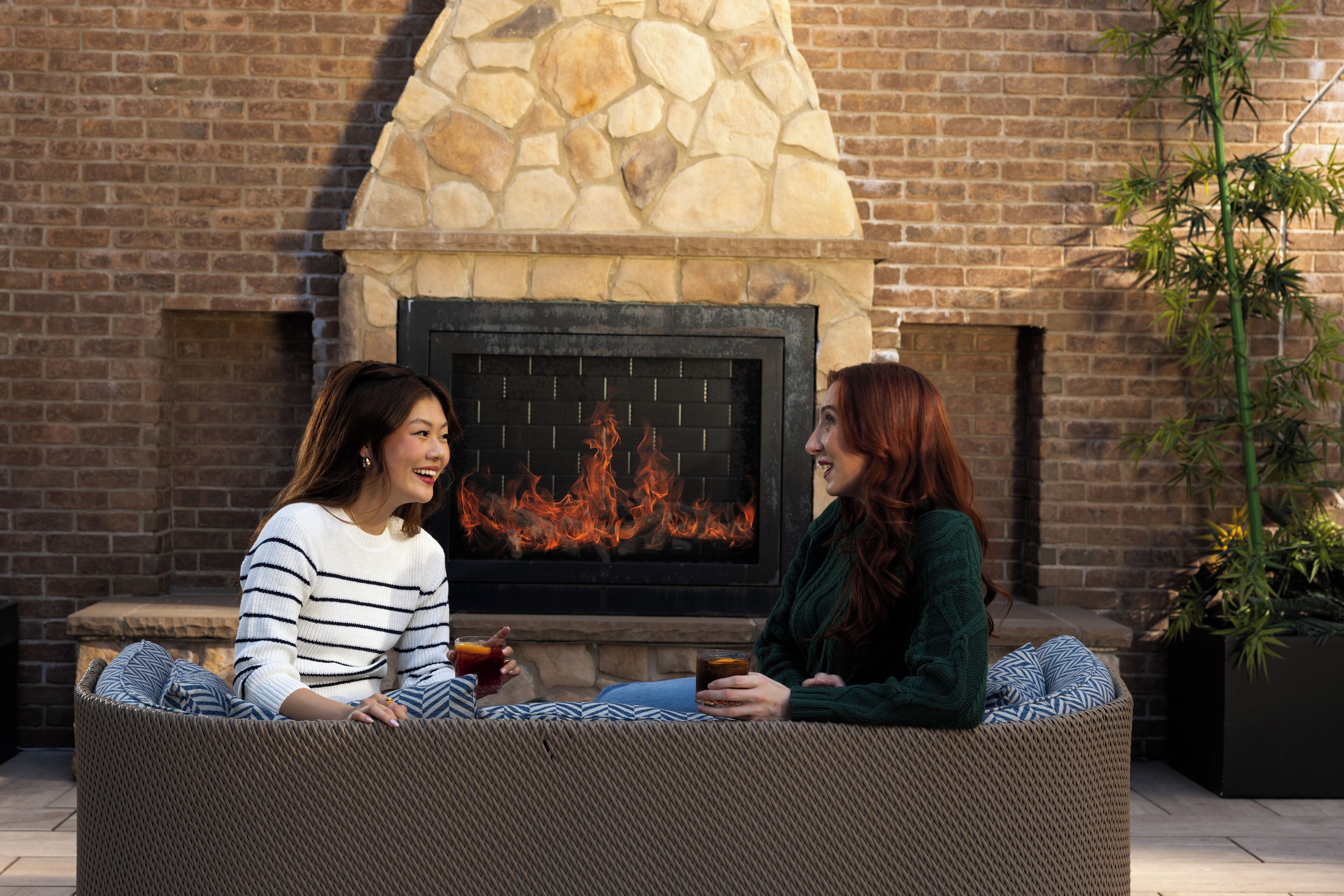 Two people sitting on wicker furniture in front of a stone fireplace with a fire.