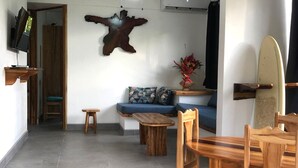 Un soggiorno con un tavolino in legno, un divano con cuscini decorati, una TV a parete e un’asse da surf appoggiata al muro.