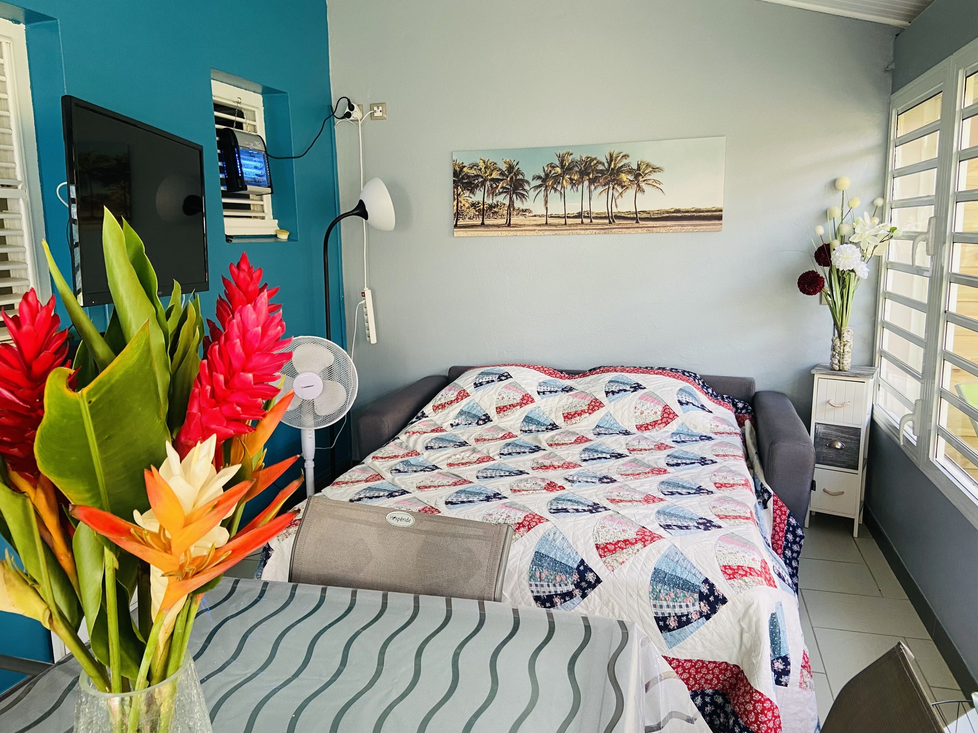 A bedroom with a bed, a desk, a chair, a standing fan, a vase with flowers, and a painting on the wall.