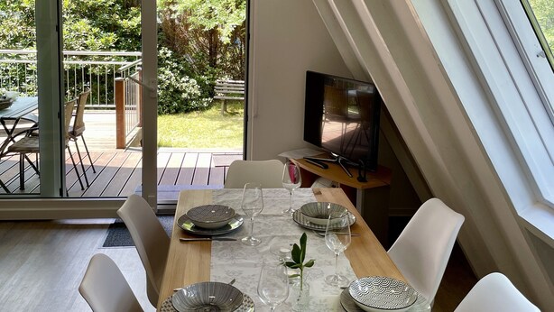 Un coin repas avec une table dressée pour quatre personnes, un téléviseur et une vue sur un jardin, accessible par une porte coulissante.