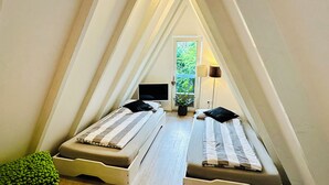Une petite chambre mansardée triangulaire, avec deux lits, une plante en pot et une fenêtre donnant sur un paysage verdoyant.