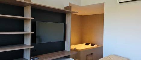 A modern hotel room with a flat-screen TV mounted on a wooden shelf, a small refrigerator, a bed with a beige duvet, and a bathroom with a bathtub and a shower.