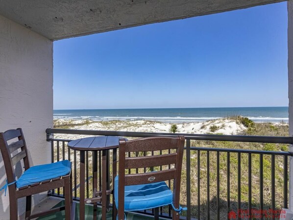 A balcony with two chairs and a table, overlooking a beach with sand dunes and the ocean.