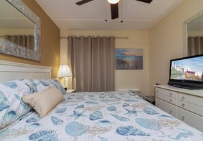 A hotel room with a bed, a television, a lamp, curtains, a painting, and a ceiling fan.