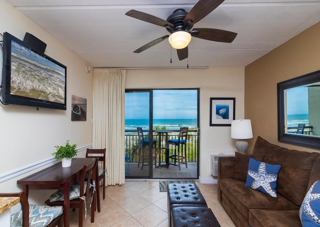 A living room with a balcony overlooking the ocean, a ceiling fan, and a television on the wall.