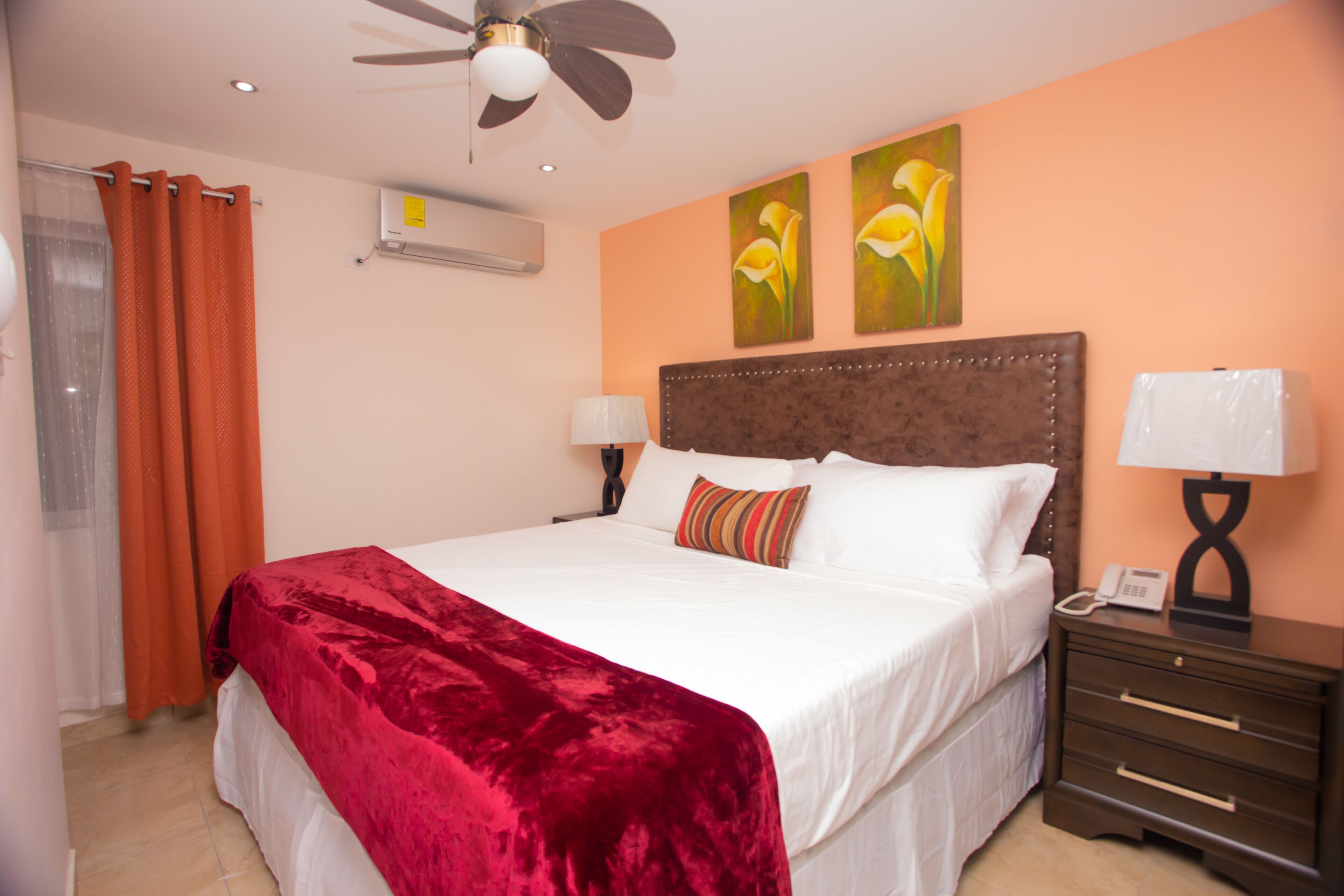 A hotel room with a large bed, two wall-mounted air conditioners, a ceiling fan, a nightstand with a lamp, and two paintings on the wall.