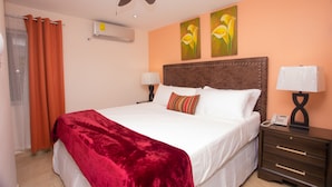 Habitación de hotel con una cama grande, dos aires acondicionados instalados en la pared, un ventilador de techo, una mesita de noche con lámpara y dos cuadros en la pared.