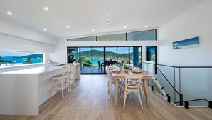 A modern kitchen with a large island, dining area with wooden chairs, and a view of the sea through large windows. - Airlie Bay View (Airlie Beach)