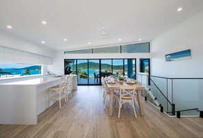 A modern kitchen with a large island, dining area with wooden chairs, and a view of the sea through large windows. - Airlie Bay View (Airlie Beach)