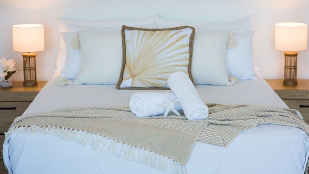 A neatly made bed with white linens, a decorative pillow, and a rolled towel. There are two bedside lamps on either side of the bed. - Airlie Bay View (Airlie Beach)