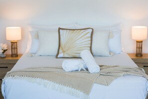 Un lit bien fait, avec du linge de lit blanc, un coussin décoratif et une serviette enroulée. Il y a deux lampes de chevet, une de chaque côté du lit.
