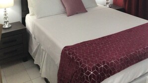 Un letto ben rifatto con cuscini bianchi e viola, una testiera scura e una tenda rossa. Sul muro sopra il letto, un quadro di un fiore, e si vede una ventola a soffitto.
