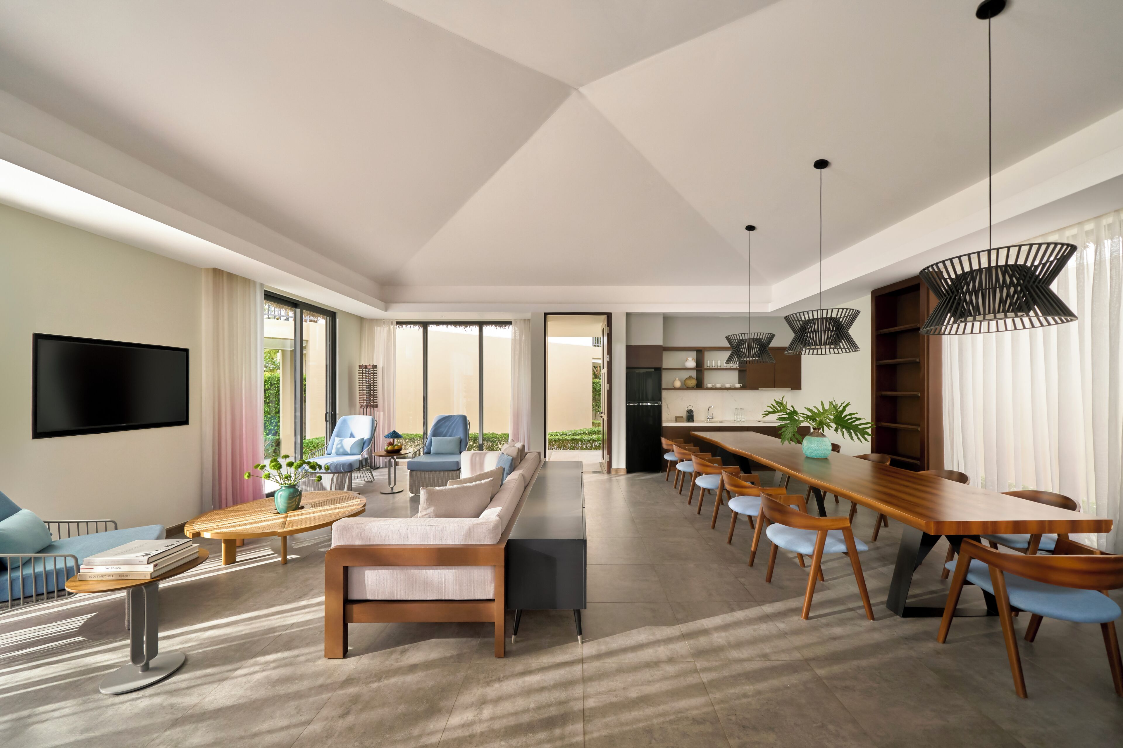 A modern living room with a sofa, coffee table, and TV. There is a dining area with a long table and chairs.