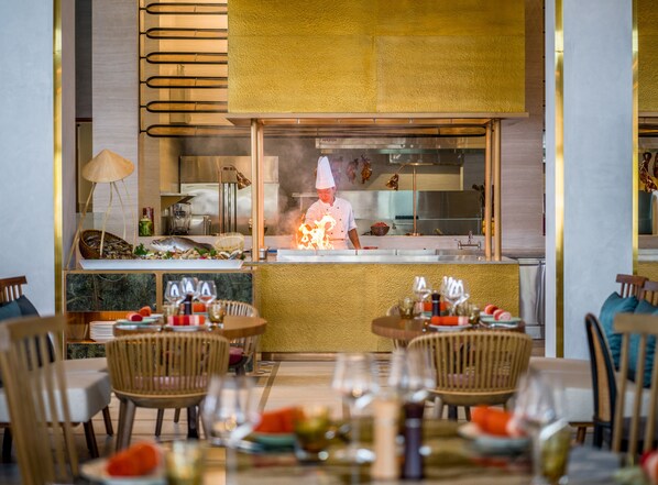 Un restaurante con un chef cocinando en la barra, sillas de madera y una mesa de mármol con copas de vino.