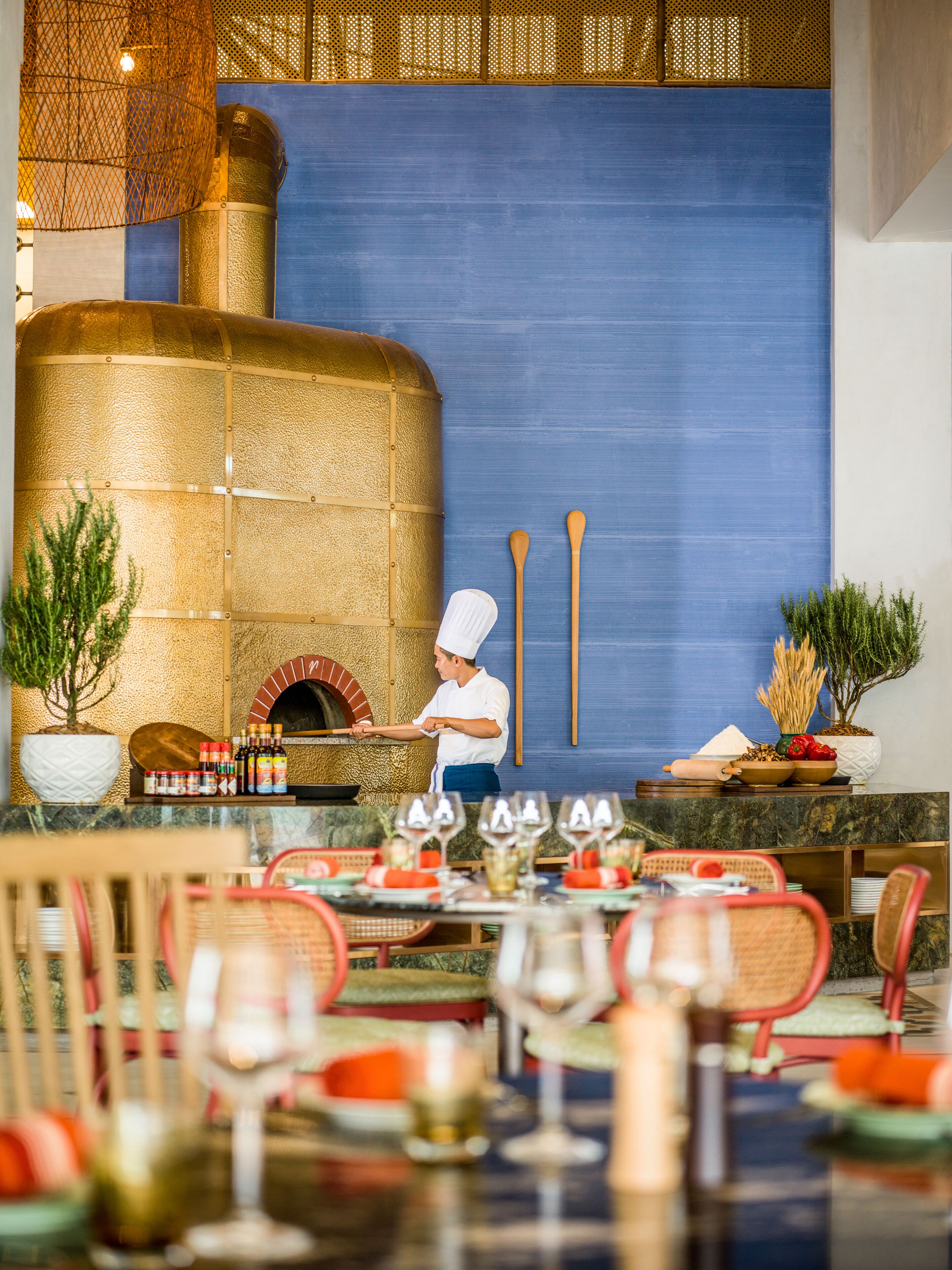 Un restaurante con un chef preparando comida, un horno de ladrillo y una zona de comedor con mesas y sillas.