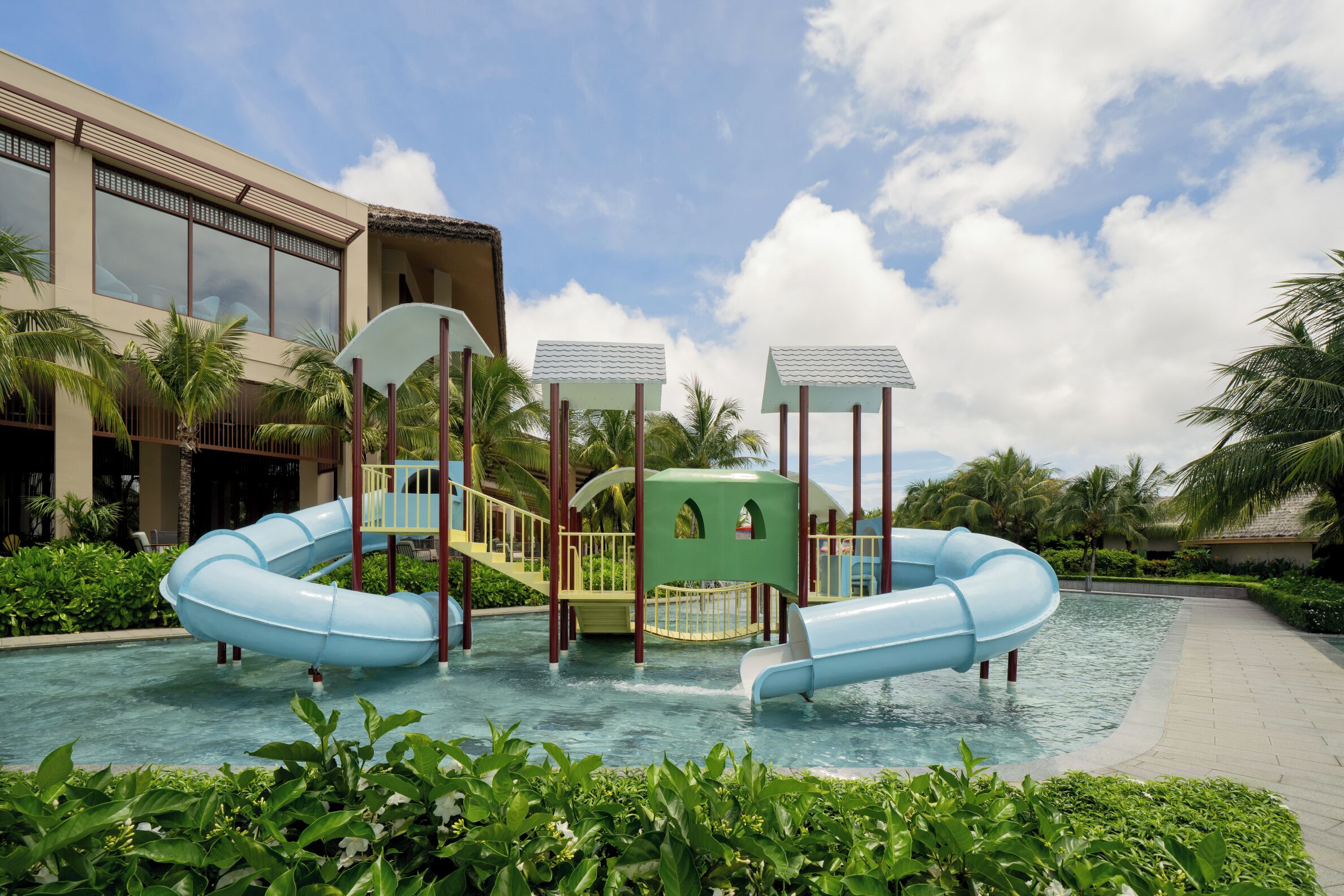 A pool area with two blue slides, a small bridge, and a building with large windows.