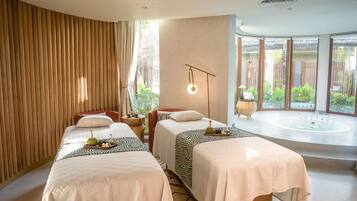 A hotel room with two beds, a large window, a bathtub, and a small table with a bottle and a bowl.