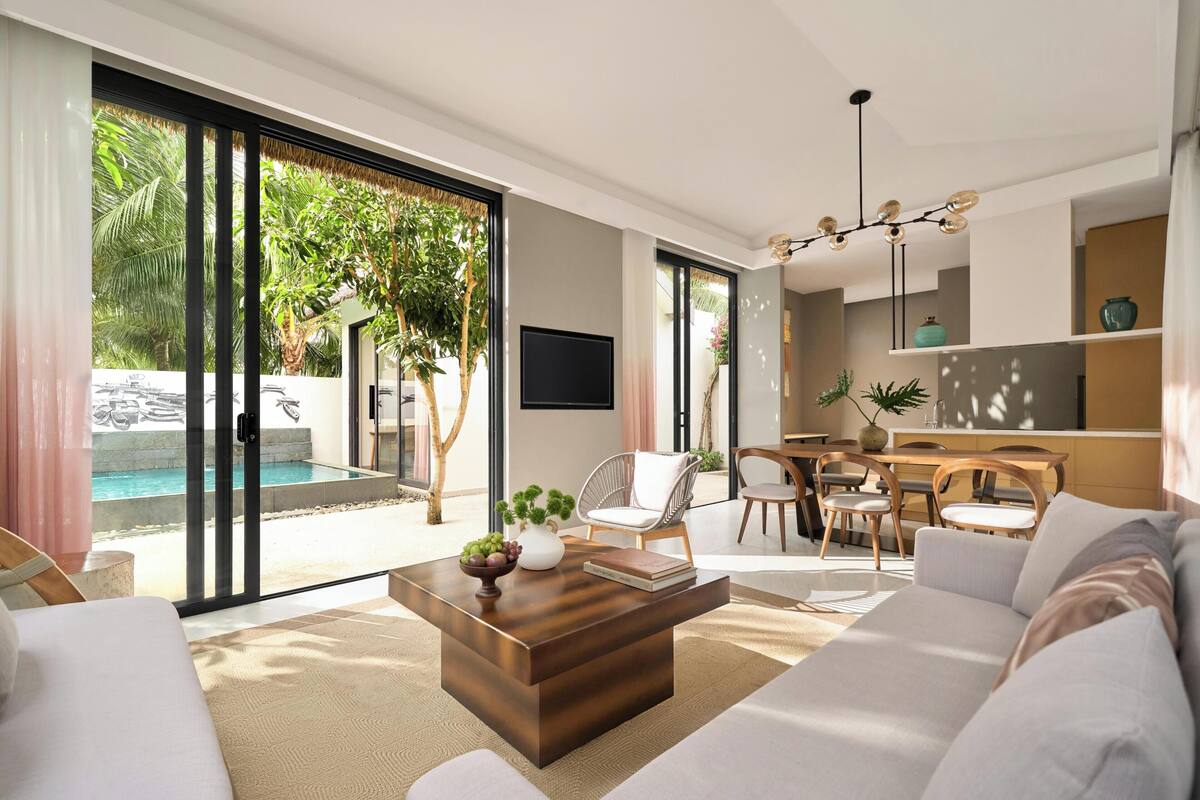 A modern living room with a sofa, coffee table, and dining area. There is a large sliding glass door leading to a pool and outdoor area.