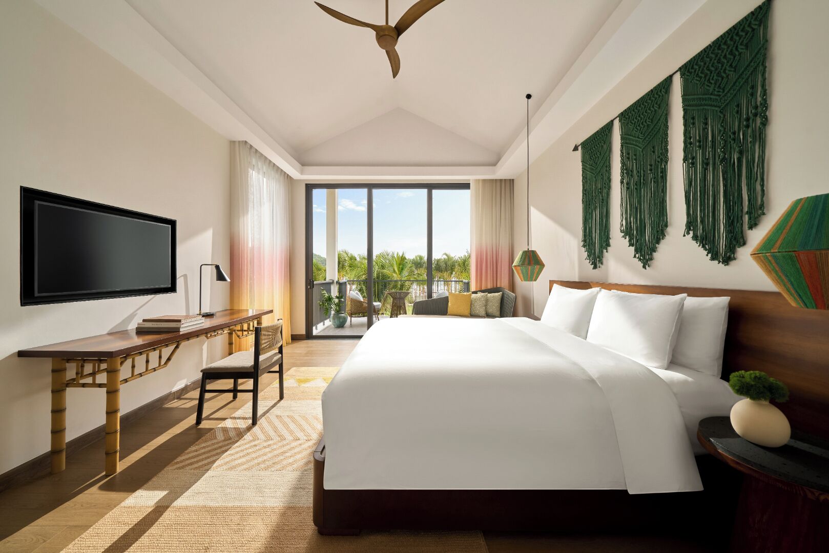 A modern hotel room with a large bed, a desk, a TV, and a balcony with a view of palm trees.