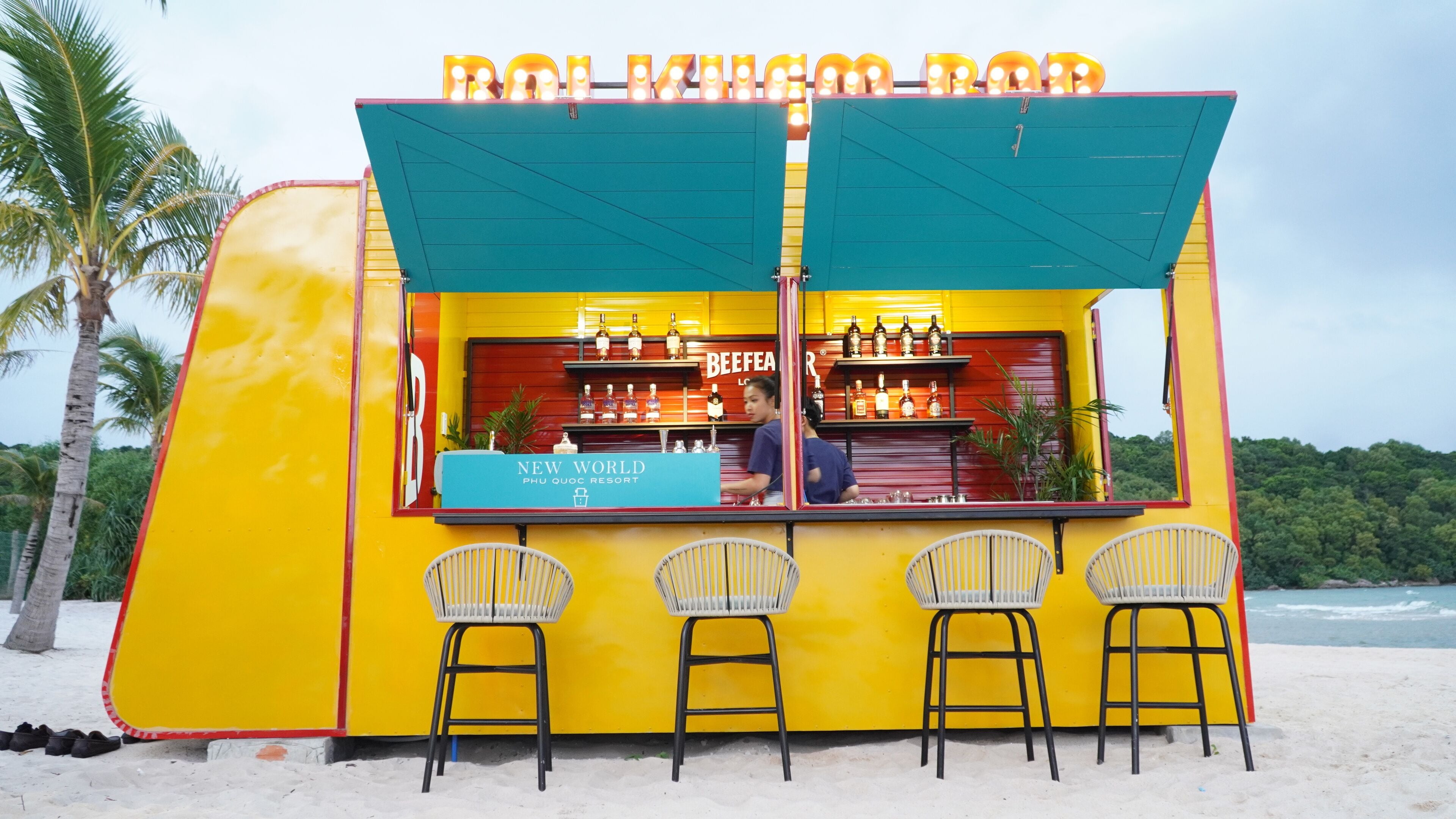 A beach bar with a bright yellow exterior, a blue sign reading "New World Phu Quoc Resort", and three white wicker chairs in front.